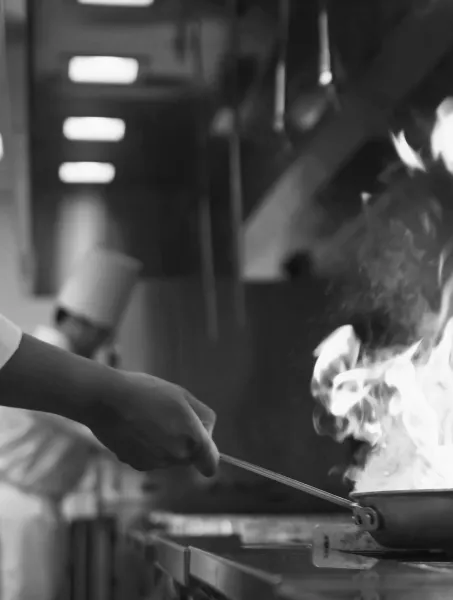 A chef holds a pan with high flames in a busy restaurant kitchen, conveying a dynamic, intense atmosphere. Another chef with a hat works in the background.