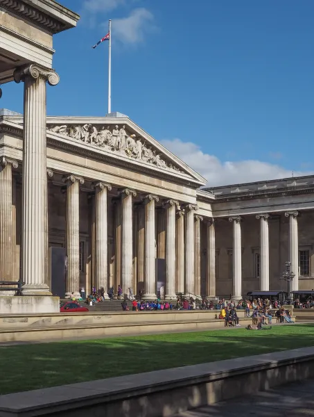 Majestic neoclassical museum building with tall columns and a detailed frieze. Visitors relax on the lawn under a clear blue sky, creating a tranquil atmosphere.