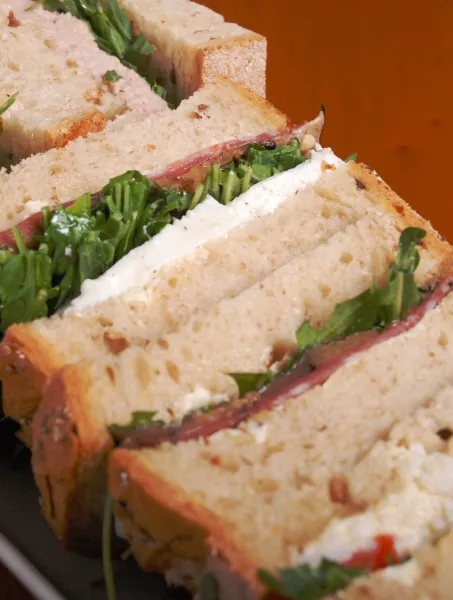 Slices of sandwiches with brown bread, arugula, and various fillings like cheese and meat are neatly arranged on a white plate over a wooden table.