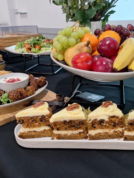 A buffet spread featuring carrot cake slices with pecans, fresh fruits like grapes, bananas, and apples, falafel with dip, and salad on a black tablecloth.
