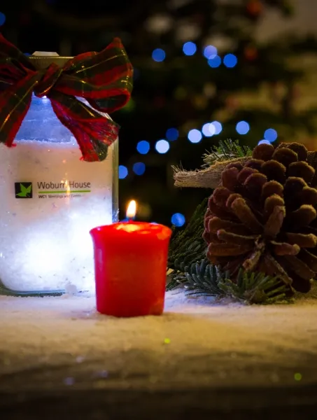 A festive scene with a glowing jar adorned with a red plaid bow next to a lit red candle. A pinecone and evergreen sprig add a rustic touch, with blurred lights in the background for a cozy holiday vibe.