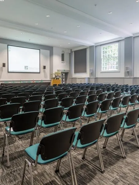 A spacious, well-lit conference room with rows of black and teal chairs facing a large projector screen. The room has tall windows and a modern, neutral decor.