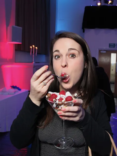 A woman enjoying a dessert in a glass, eating with a spoon. The room has pink and blue lighting, creating a festive atmosphere with a candelabrum in the background.