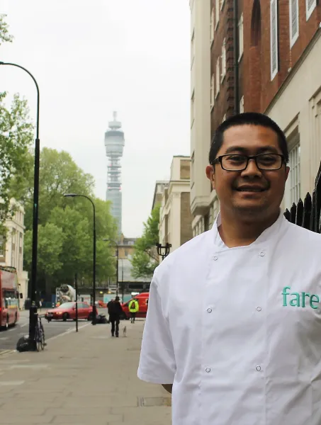 Chef in a white uniform, smiling on a city street with trees and buildings. A distant telecom tower rises in the background, conveying a professional yet relaxed atmosphere.
