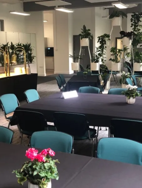 Spacious meeting room with black-tablecloth tables and teal chairs, decorated with potted plants. Bright, modern ambiance with ample natural light.