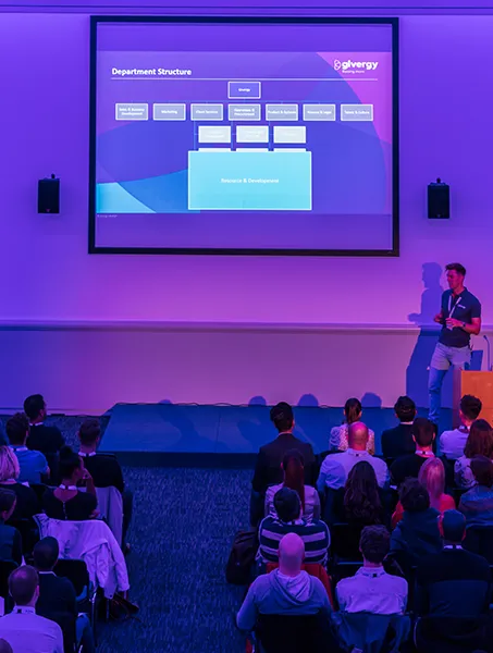 A speaker presents a departmental structure slide to an audience in a dimly lit conference room with blue and purple lighting.