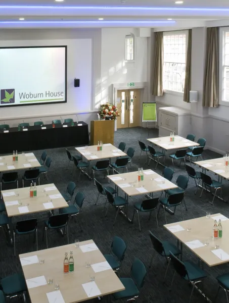 Spacious conference room with arranged tables, teal chairs, and a podium. A large screen displays "Woburn House." Bright and professional atmosphere.