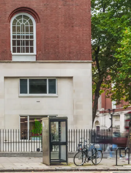 Street view of Woburn House, a red-brick building with large windows, and a blurred red double-decker bus passing by. Bicycles and a tree are visible.