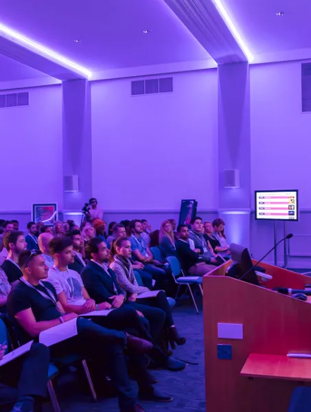 A speaker presents to an attentive audience in a purple-lit conference room. The mood is focused, with a screen displaying a presentation slide.