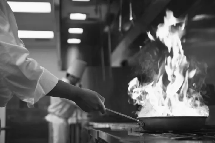 A chef holds a pan with high flames in a busy restaurant kitchen, conveying a dynamic, intense atmosphere. Another chef with a hat works in the background.