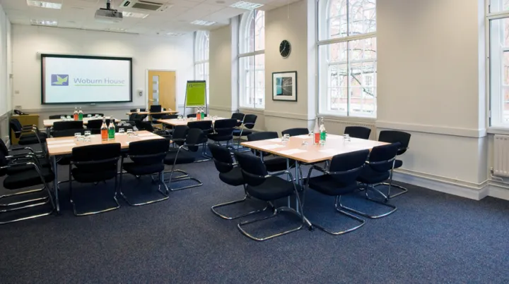 A conference room with multiple tables and black chairs, each table set with water bottles and glasses. A projection screen reads "Woburn House."