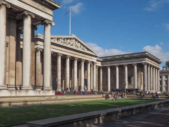 Majestic neoclassical museum building with tall columns and a detailed frieze. Visitors relax on the lawn under a clear blue sky, creating a tranquil atmosphere.