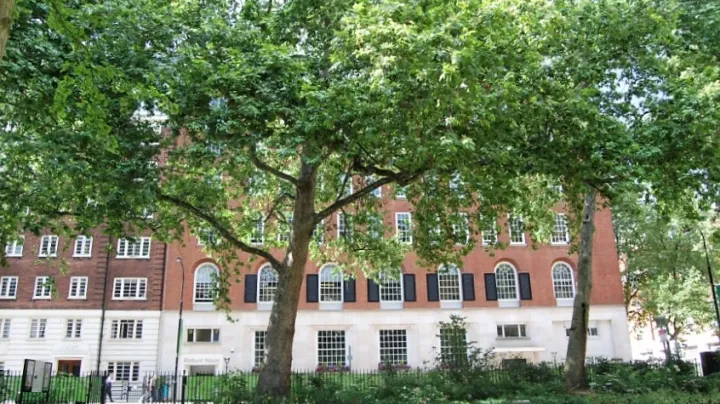 A red brick building with large windows is partially obscured by lush green trees in the foreground. The scene feels serene and summery.