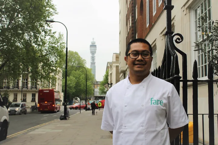 Chef in a white uniform, smiling on a city street with trees and buildings. A distant telecom tower rises in the background, conveying a professional yet relaxed atmosphere.