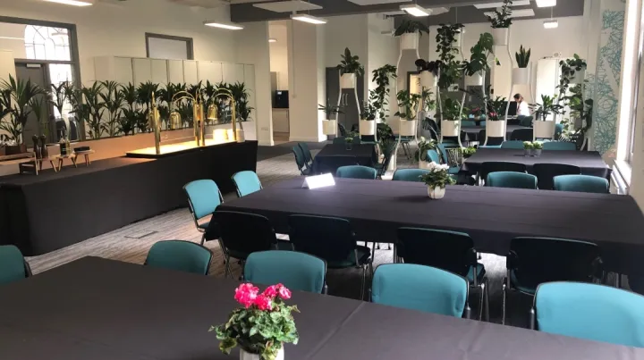Spacious meeting room with black-tablecloth tables and teal chairs, decorated with potted plants. Bright, modern ambiance with ample natural light.