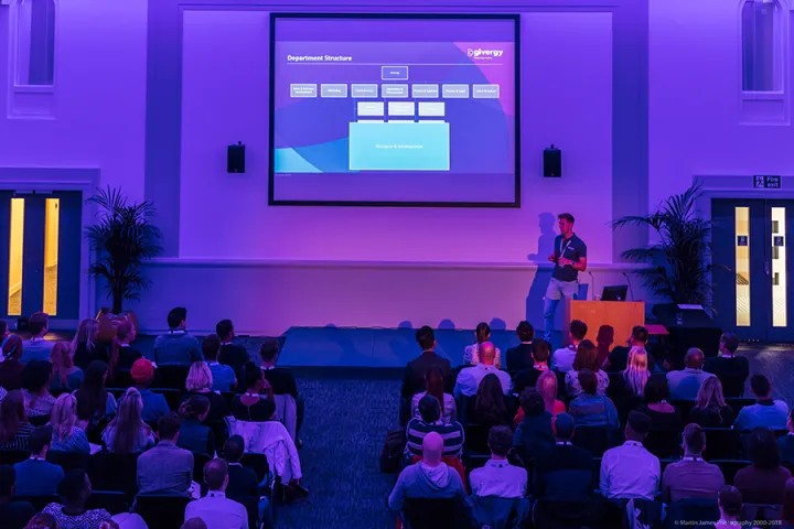 A speaker presents a departmental structure slide to an audience in a dimly lit conference room with blue and purple lighting.