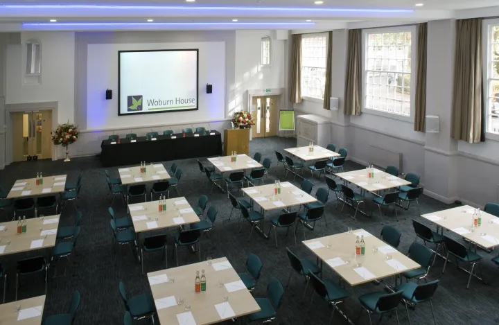 Spacious conference room with arranged tables, teal chairs, and a podium. A large screen displays "Woburn House." Bright and professional atmosphere.