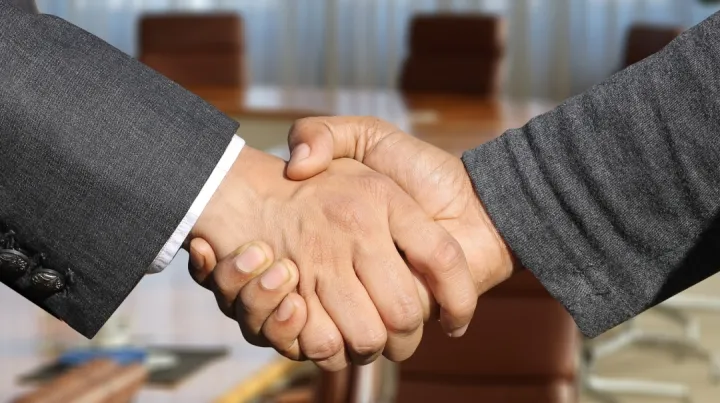 Two hands in business suits shake hands in a boardroom setting, symbolizing agreement or partnership. The mood is professional and cooperative.
