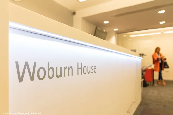 Lobby of Woburn House with a modern, white reception desk prominently displaying the name. A person stands in the blurred background, conveying a formal atmosphere.