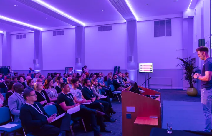 A speaker presents to an attentive audience in a purple-lit conference room. The mood is focused, with a screen displaying a presentation slide.